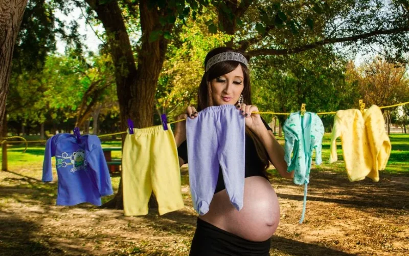 Expecting mother hanging colorful baby clothes on a line in a serene outdoor setting.