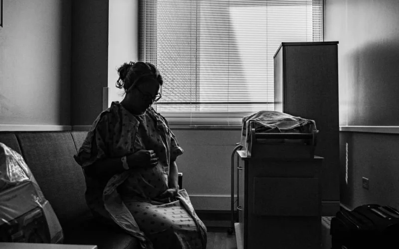 A thoughtful pregnant woman in a hospital room, captured in black and white.
