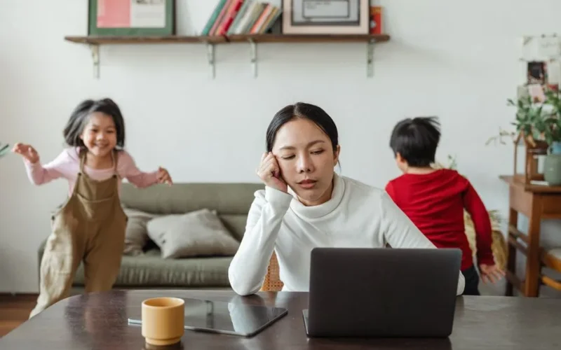 Exhausted young Asian female working distantly from home with happy noisy children playing around