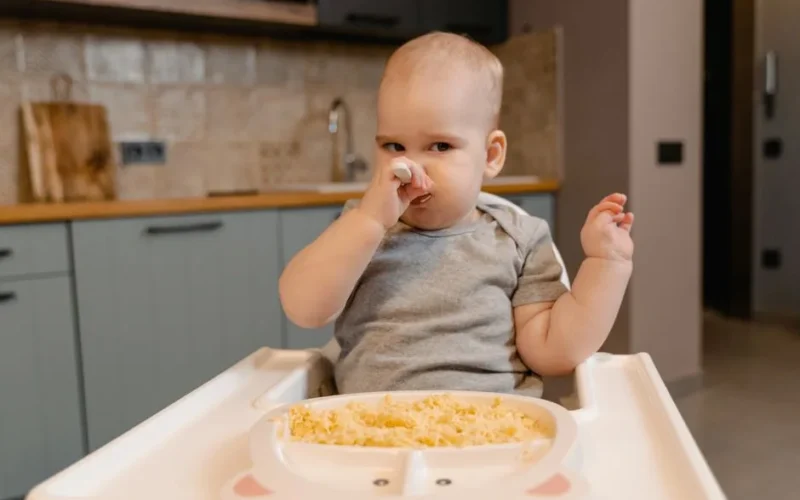 Adorable baby in onesie feeding themselves in a high chair indoors.