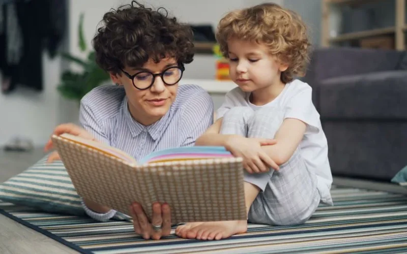 Mother and young son lying on carpet, engaged in reading a book together happily.