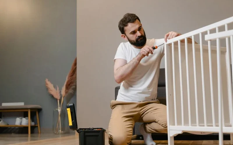 Father kneels to fix a white baby crib using a screwdriver in a contemporary nursery setting.