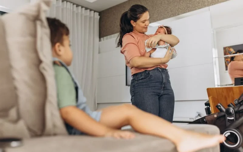 A mother lovingly holds her newborn while a toddler sits on the couch in a cozy living room setting.