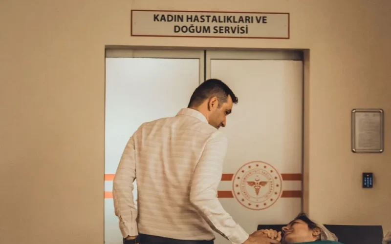 A man supports a woman in the maternity ward of a hospital, holding hands.