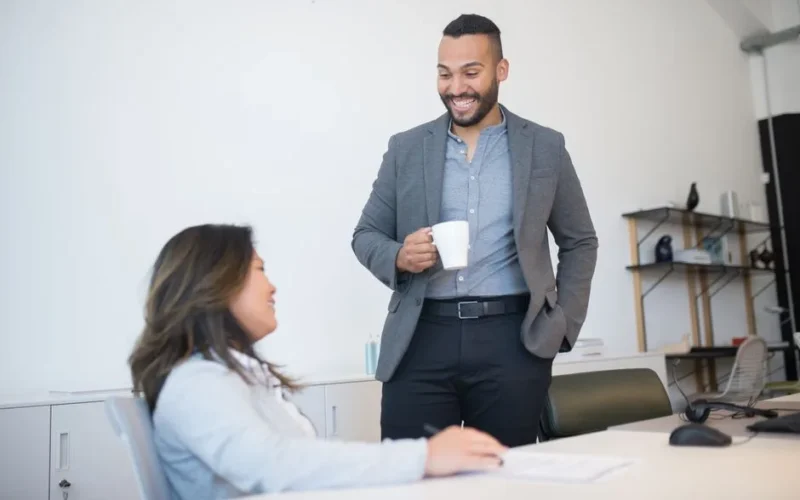 Two business professionals smiling and talking during a coffee break in a modern office setting.