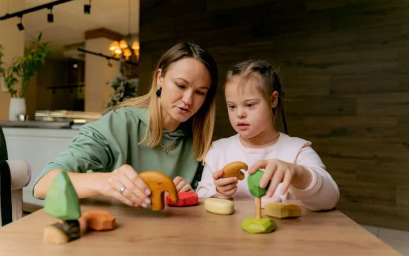 A mother and daughter engaging in play with wooden toys at a table indoors.