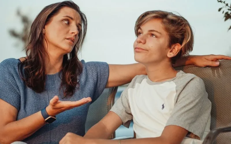 A mother and her teenage son engage in a conversation sitting outside, expressing familial warmth and connection.