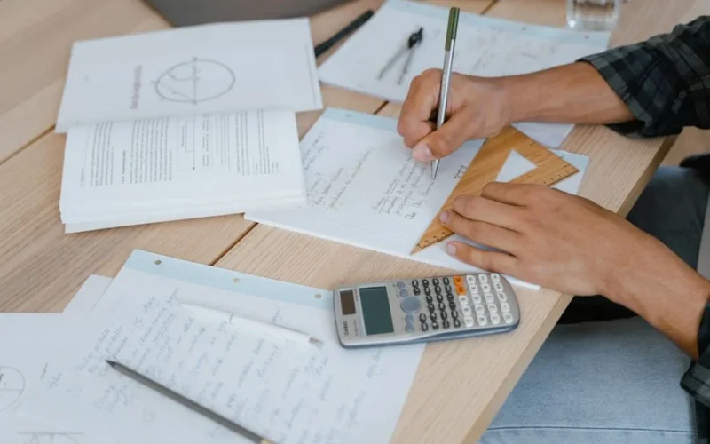 A student engaged in solving math problems using a calculator and stationery on a desk.