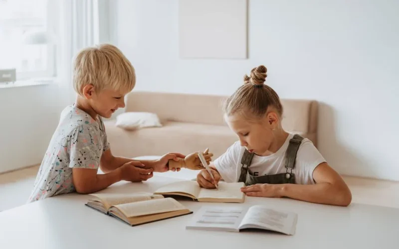 Blonde boy and girl studying together indoors, focusing on writing and learning.