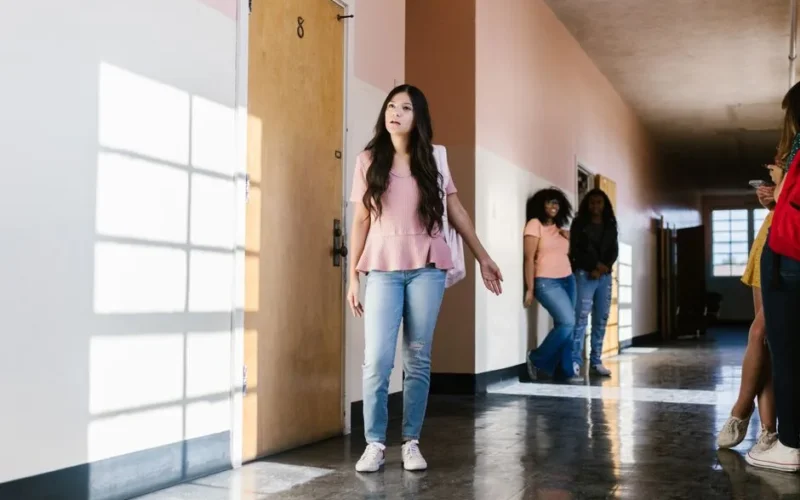 Teenage girl in school corridor looking thoughtful, surrounded by peers, captured in natural lighting.