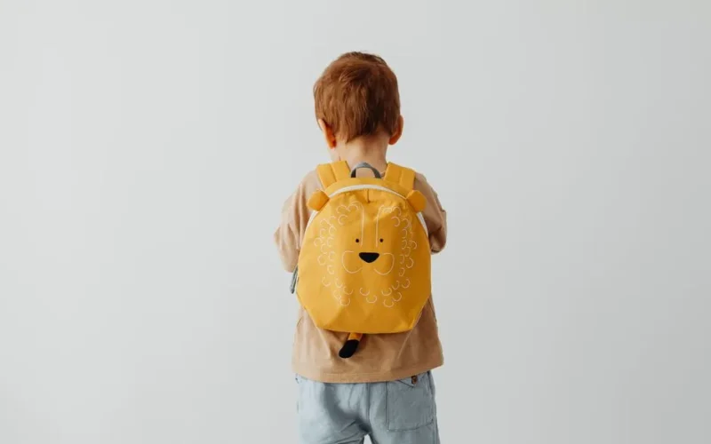 Cute child with red hair wearing a playful lion backpack facing away.