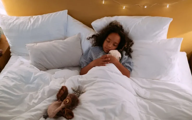 A young girl sleeps comfortably in bed hugging a plush toy, surrounded by pillows and soft lighting.