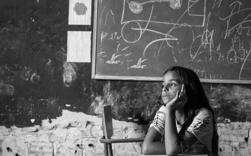 Black and white photo of a young girl sitting thoughtfully in a classroom in Bezerros, Brazil.
