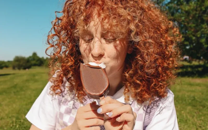 Curly-haired woman savoring a chocolate ice cream bar in a sunny outdoor park.