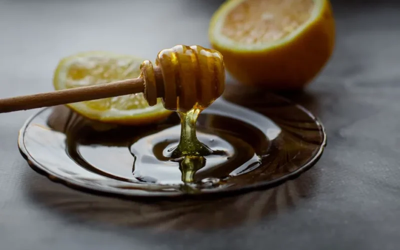 Delicious honey flowing down from dipper into saucer with lemon slice placed on black table