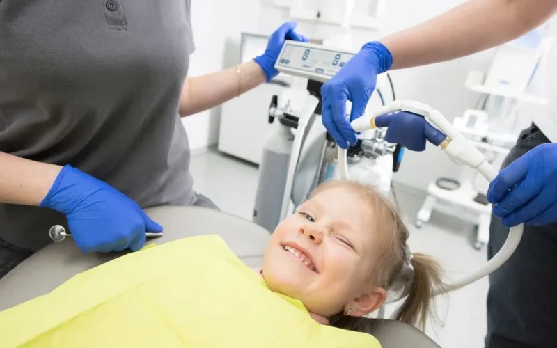 A cheerful child winks while sitting in a dental chair, surrounded by dental professionals.