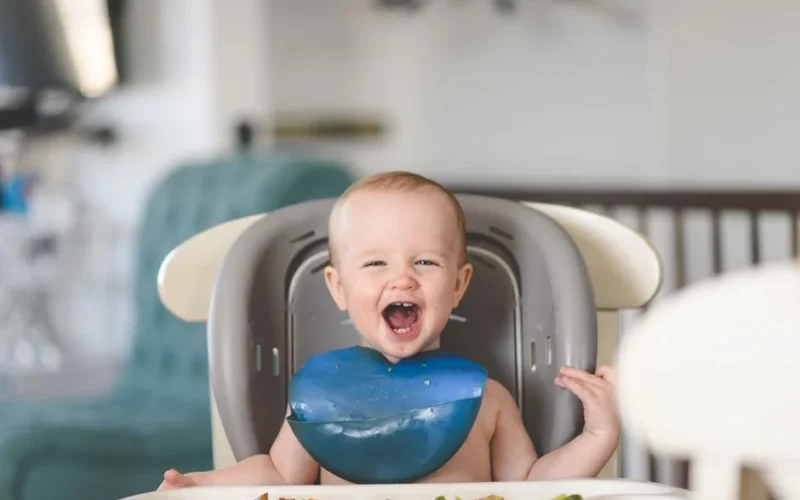 Happy baby laughing in a high chair during mealtime, wearing a blue bib.