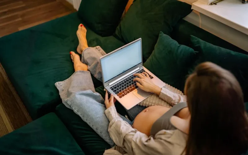 Pregnant woman sitting comfortably on a sofa using a laptop, working remotely.
