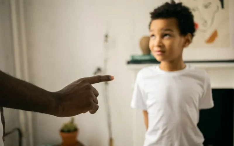 Crop unrecognizable strict black man scolding and disciplining guilty attentive little son standing with hands behind back and looking up