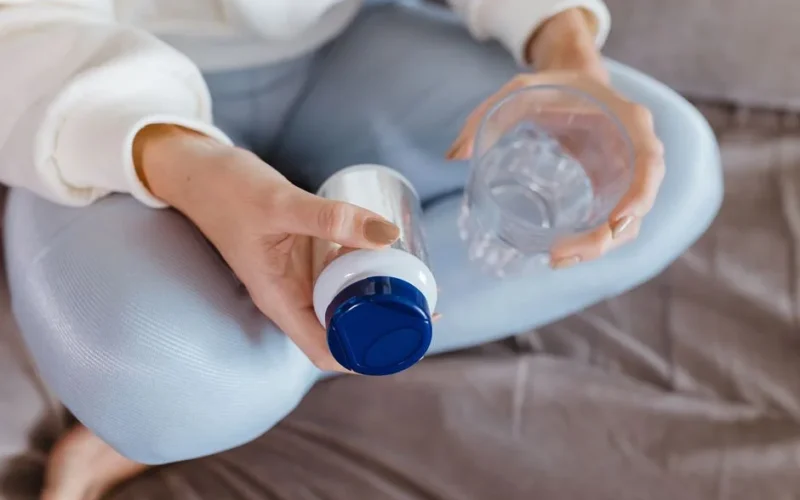 Close-up of a woman sitting cross-legged, holding a pill bottle and a glass of water.