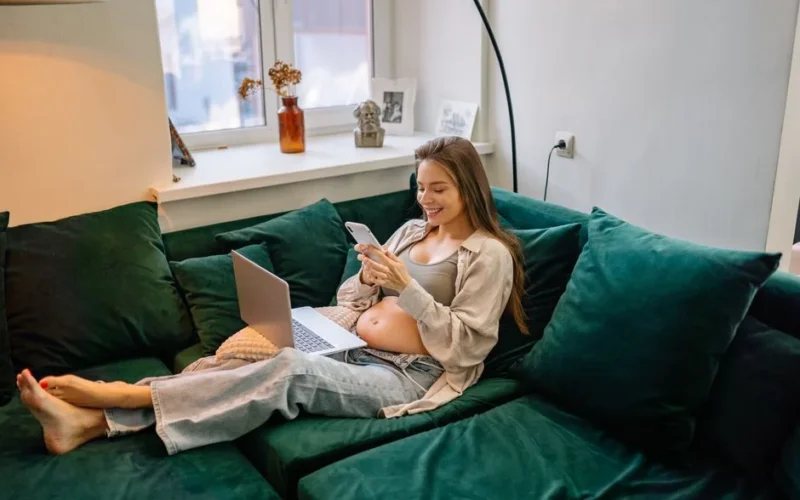 Pregnant woman lounging on a sofa, using a smartphone, and working on a laptop indoors.