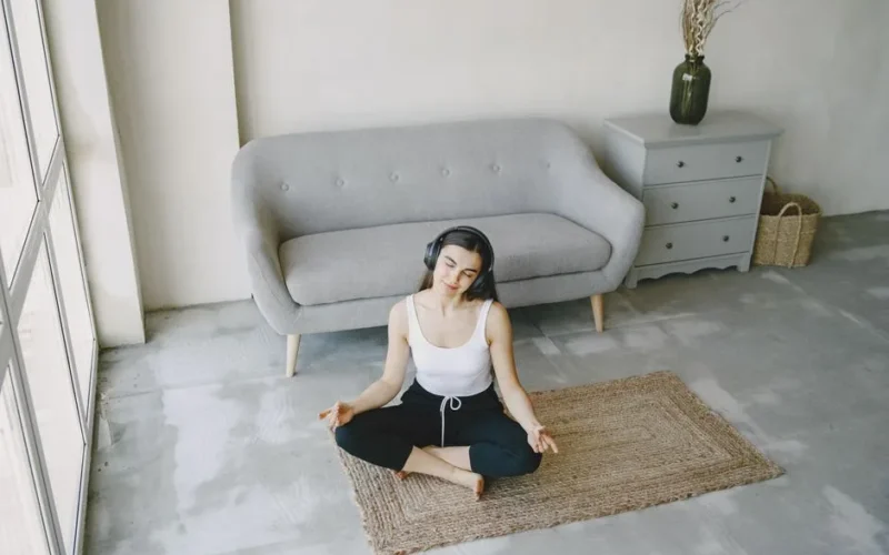 A young woman sits on a mat in a contemporary living room, meditating with headphones on.