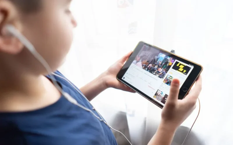 Child holding a tablet indoors, watching videos with headphones, showcasing technology usage by kids.