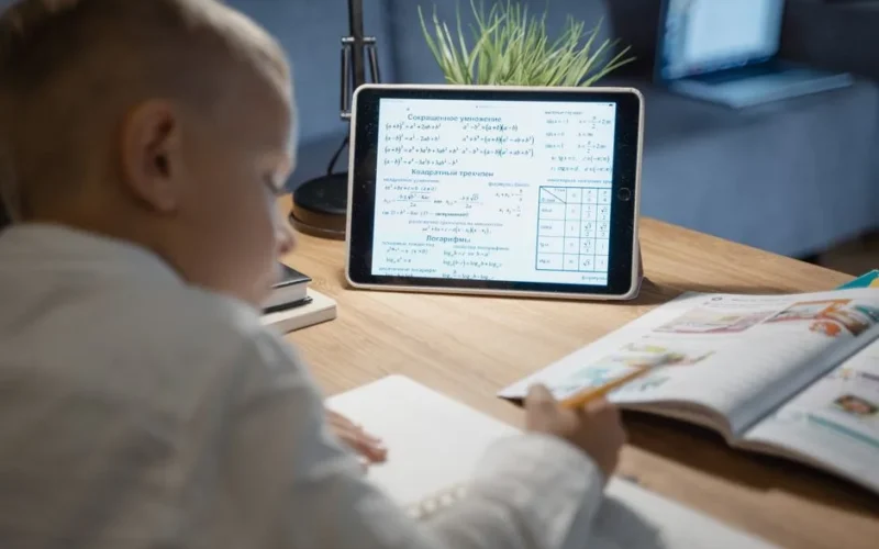 A young boy focuses on math problems on a tablet while studying at a desk indoors.