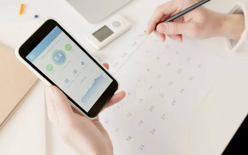 Close-up of hands using a smartphone to track health stats while planning on a calendar.