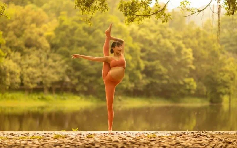 Pregnant woman in orange activewear practicing yoga by a serene lake outdoors.