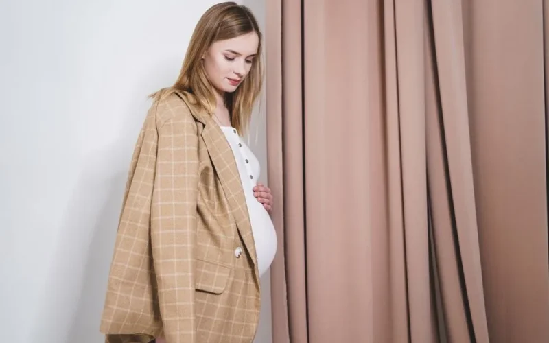 Pregnant woman in a stylish tan blazer looking down indoors, showcasing elegance and maternity fashion.