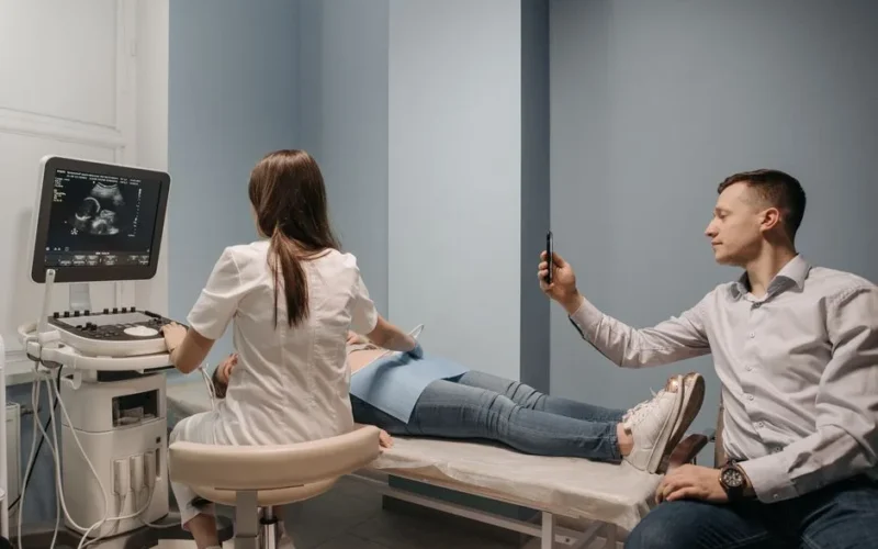 A doctor performs an ultrasound on a pregnant woman, while a man takes a photo in a clinical environment.