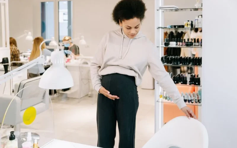 Pregnant woman with afro hair inspects a white chair in a modern salon setting.