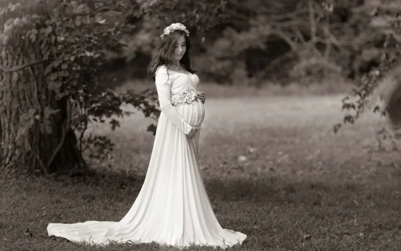 Monochrome portrait of an expectant woman in a flowing dress in a serene outdoor setting.
