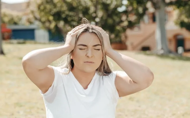 Adult woman holding her head in pain, sitting outdoors under sunlight, likely experiencing a migraine.
