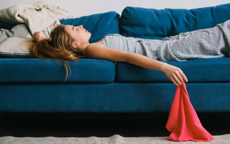 Side view of young exhausted woman with rag lying on couch in living room in daylight