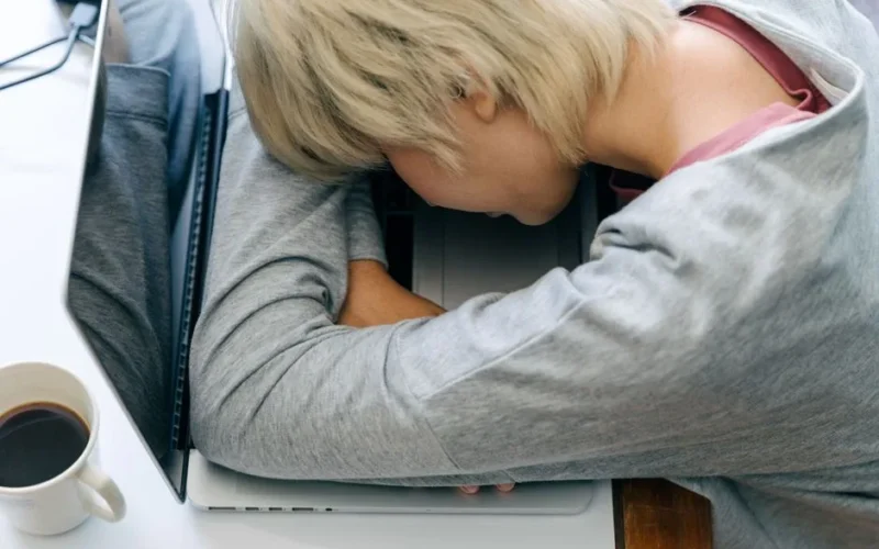 From above of anonymous female in casual clothes resting head on netbook keyboard near cup of coffee after long working day