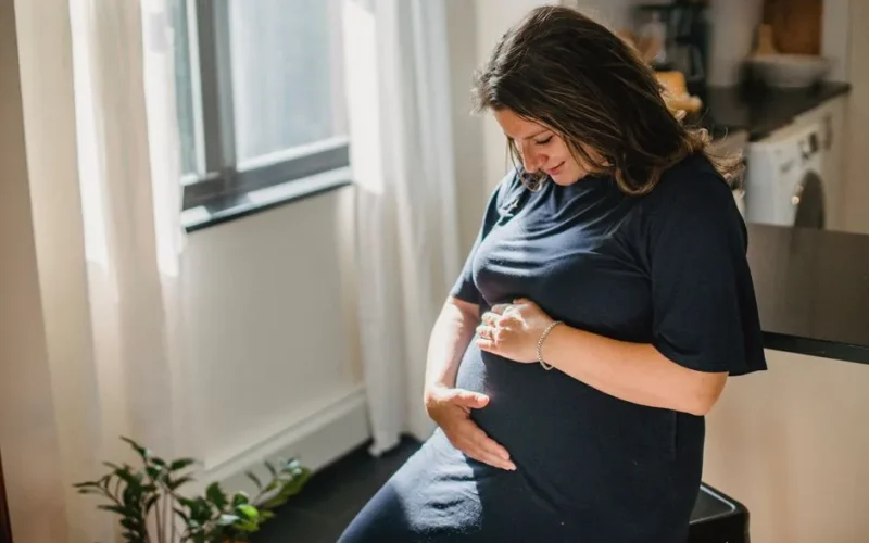 Positive pregnant female with brown hair in dress sitting on chair and touching belly in cozy apartment in daytime
