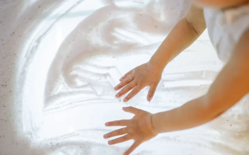 A child happily plays with foamy substance using hands on a light surface.