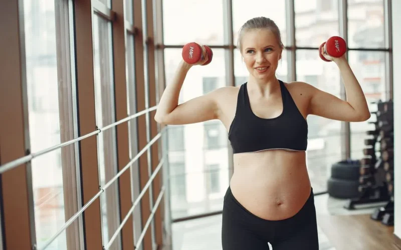 Expectant woman in workout gear lifts dumbbells indoors, embodying fitness and health.